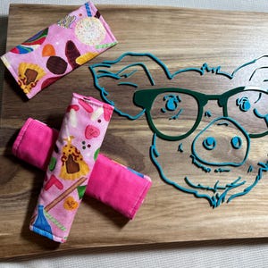 Puede incluir: Tres agarraderas de olla de tela rosa con un diseño colorido de caramelos y dulces. Las agarraderas están sobre una tabla de cortar de madera con un diseño de cerdo turquesa.