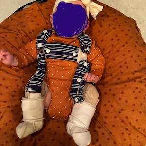 May include: A baby wearing a brown and white polka dot onesie with a blue and white striped bib and a white headband with a bow. The baby is lying on a brown and white patterned pillow.