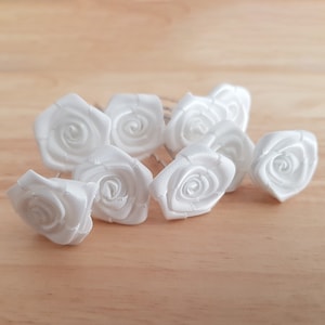 Può includere: Un set di otto forcine per capelli in raso bianco a forma di rosa. Le forcine sono disposte in un gruppo su una superficie di legno marrone chiaro.