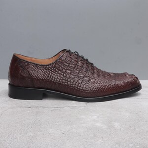 Side view of a single dark brown men’s wholecut shoe with Alligator, closed lacing system, low stacked heel, and leather outsole, placed on light grey concrete surface.