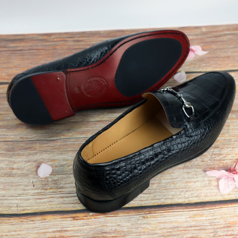 May include: A pair of black leather loafers with a crocodile pattern. The loafers have a silver buckle detail and a red leather insole.