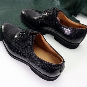 K&ouml;nnte beinhalten: Ein Paar schwarze Lederschuhe mit Kroko-Muster. Die Schuhe haben Schn&uuml;rsenkel und eine abgerundete Spitze.