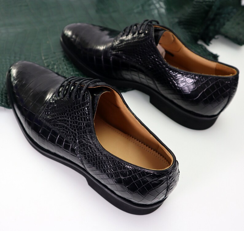 K&ouml;nnte beinhalten: Ein Paar schwarze Lederschuhe mit Kroko-Muster. Die Schuhe haben Schn&uuml;rsenkel und eine abgerundete Spitze.