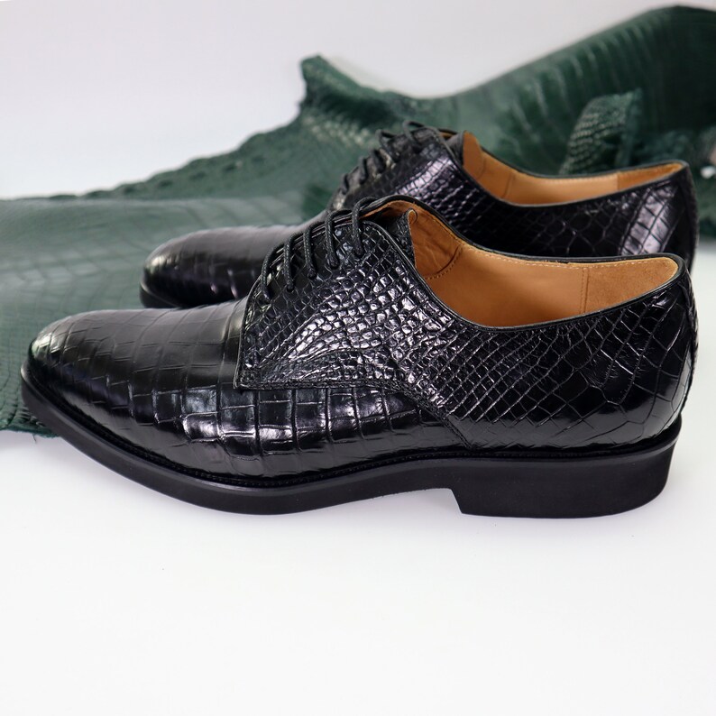 K&ouml;nnte beinhalten: Ein Paar schwarze Lederschuhe mit Kroko-Muster. Die Schuhe haben einen Schn&uuml;rverschluss und einen niedrigen Absatz.