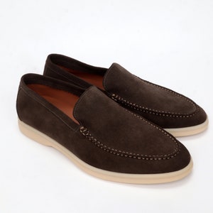 Op de afbeelding: Een paar bruine su&egrave;de loafers met een witte rubberen zool. De loafers hebben een gestikt detail rond de rand van de schoen.