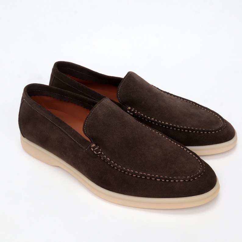 Op de afbeelding: Een paar bruine su&egrave;de loafers met een witte rubberen zool. De loafers hebben een gestikt detail rond de rand van de schoen.