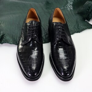 K&ouml;nnte beinhalten: Ein Paar schwarze Lederschuhe mit Kroko-Muster. Die Schuhe haben Schn&uuml;rsenkel und spitze Zehen.