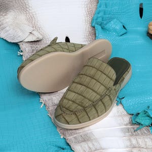 May include: Two olive green leather slip-on shoes with a textured, crocodile-like pattern. The shoes have a light beige sole and a dark green interior. The background features blue and white textured leather.