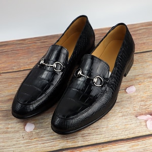 May include: A pair of black leather loafers with a crocodile pattern and silver metal hardware. The loafers have a pointed toe and a low heel.