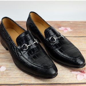 May include: A pair of black leather loafers with a crocodile pattern and silver metal buckles. The loafers have a pointed toe and a low heel.