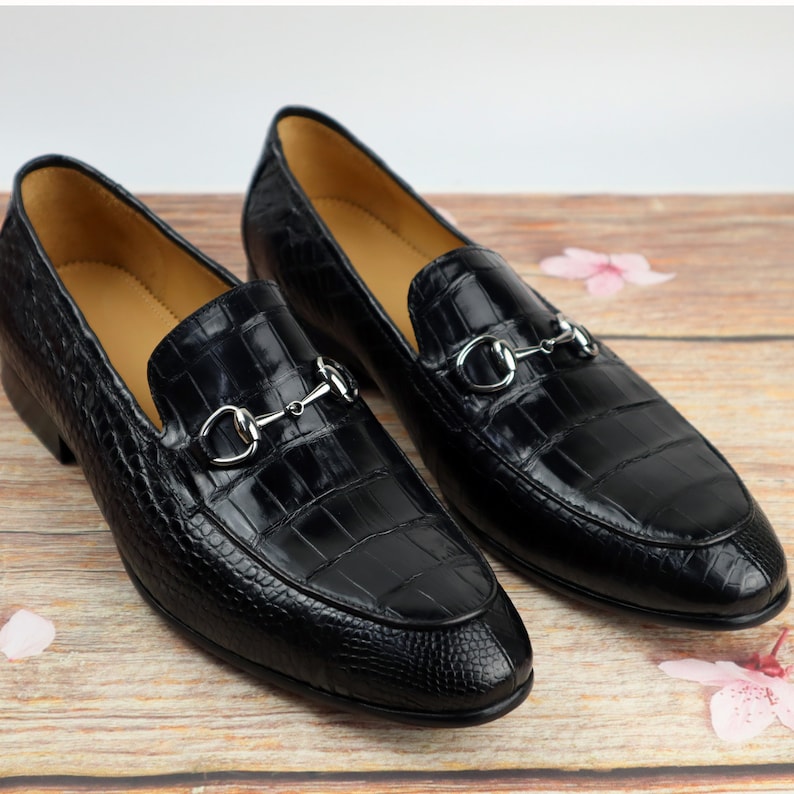 May include: A pair of black leather loafers with a crocodile pattern and silver metal buckles. The loafers have a pointed toe and a low heel.