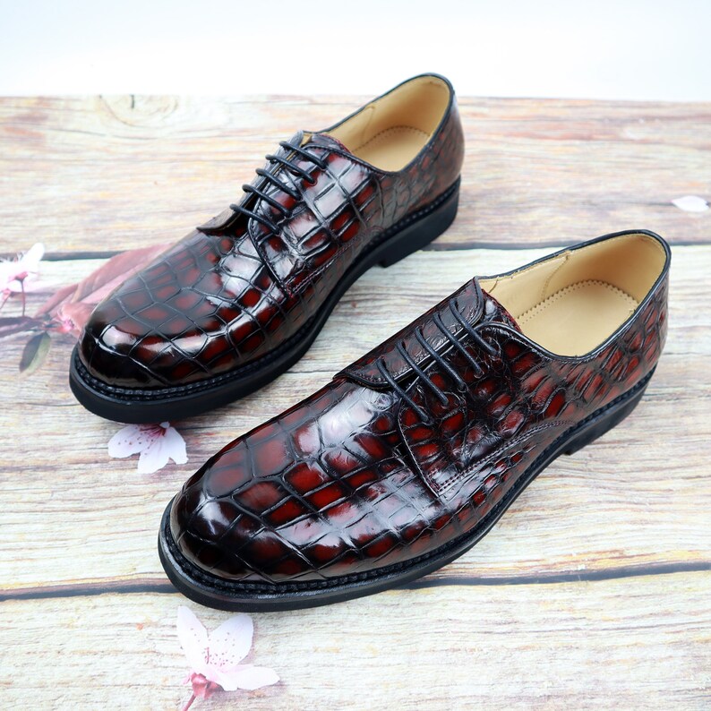 Peut inclure: Une paire de chaussures en cuir marron &agrave; lacets avec un motif crocodile. Les chaussures ont une semelle en caoutchouc noir et sont sur une surface en bois.