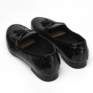 May include: A pair of black leather loafers with a crocodile skin texture. Each shoe features a decorative tassel and a low heel. The shoes are set against a white background, highlighting their glossy finish and classic design.