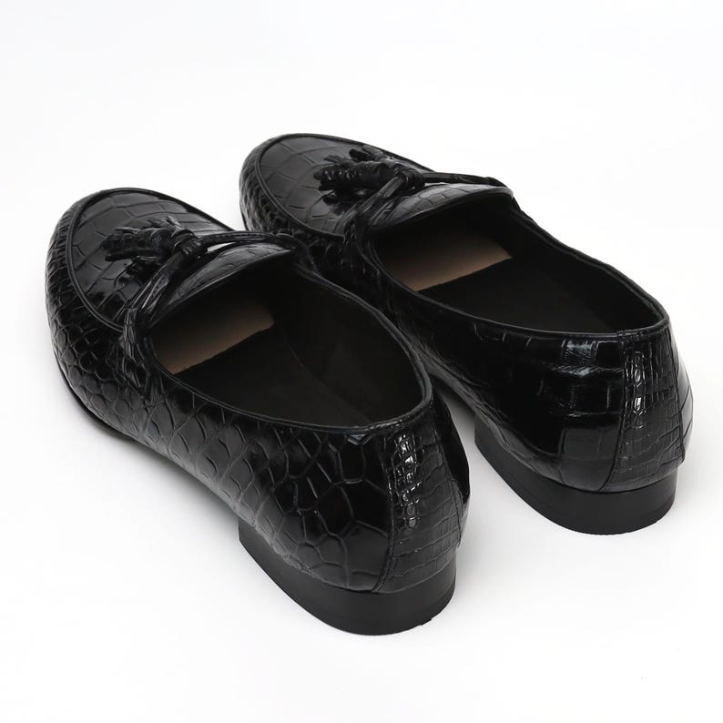 May include: A pair of black leather loafers with a crocodile skin texture. Each shoe features a decorative tassel and a low heel. The shoes are set against a white background, highlighting their glossy finish and classic design.