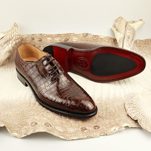 May include: A pair of brown crocodile leather lace-up shoes with a black rubber sole. The shoes are on a white background.