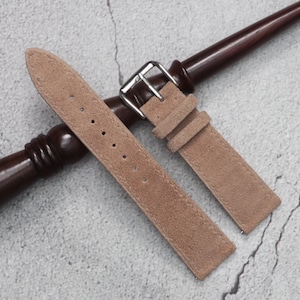 Könnte beinhalten: Ein hellbraunes Uhrenarmband aus Wildleder mit matter, texturierter Oberfläche, einer silberfarbenen Metallschnalle und zwei passenden Wildlederschlaufen. Das Armband hat eine Reihe von gestanzten Löchern zur Anpassung und liegt auf einem dunklen Holzstab.