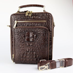 May include: A brown leather briefcase with a crocodile skin texture. The bag has a top handle, a detachable shoulder strap, and multiple zippered compartments. Gold-colored zippers and hardware provide accents.