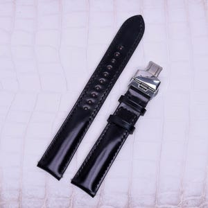 May include: A black leather watch strap featuring a polished silver-tone metal deployment clasp. The strap includes two leather loops and a series of punched holes for adjustment, showcasing a smooth, glossy finish and precise stitching along the edges.
