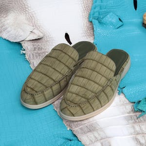 May include: A pair of olive green leather slip-on shoes with a crocodile skin pattern. The shoes have a rounded toe and a light-colored sole. The background features blue and white textured leather.