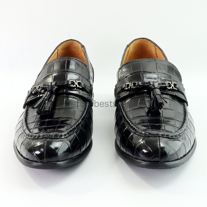 May include: A pair of black leather loafers with a crocodile pattern. The loafers feature a tassel detail and a chain accent.