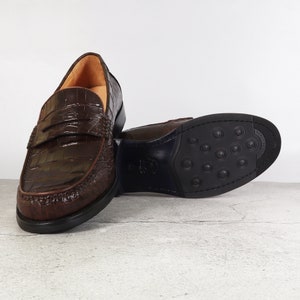 Puede incluir: Un par de mocasines de cuero marr&oacute;n con un patr&oacute;n de cocodrilo en relieve. Los mocasines tienen un dise&ntilde;o de deslizamiento y una punta redonda.