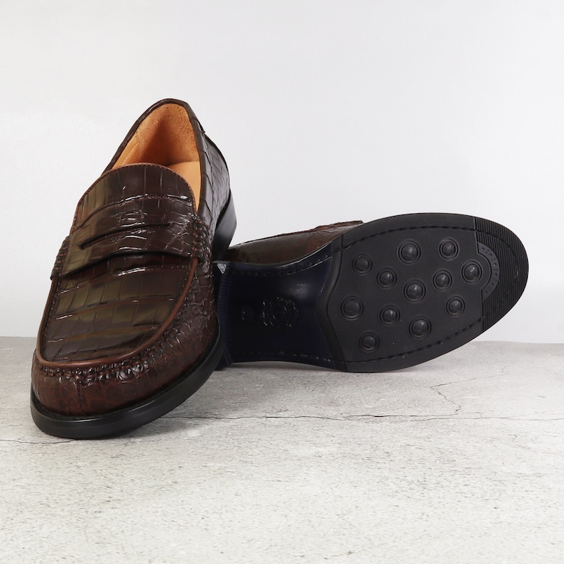 Puede incluir: Un par de mocasines de cuero marr&oacute;n con un patr&oacute;n de cocodrilo en relieve. Los mocasines tienen un dise&ntilde;o de deslizamiento y una punta redonda.