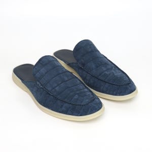 May include: A pair of blue suede slip-on shoes with a crocodile pattern. The shoes have a rounded toe and a cream-colored sole. The interior is a darker blue. The shoes are displayed on a white background.