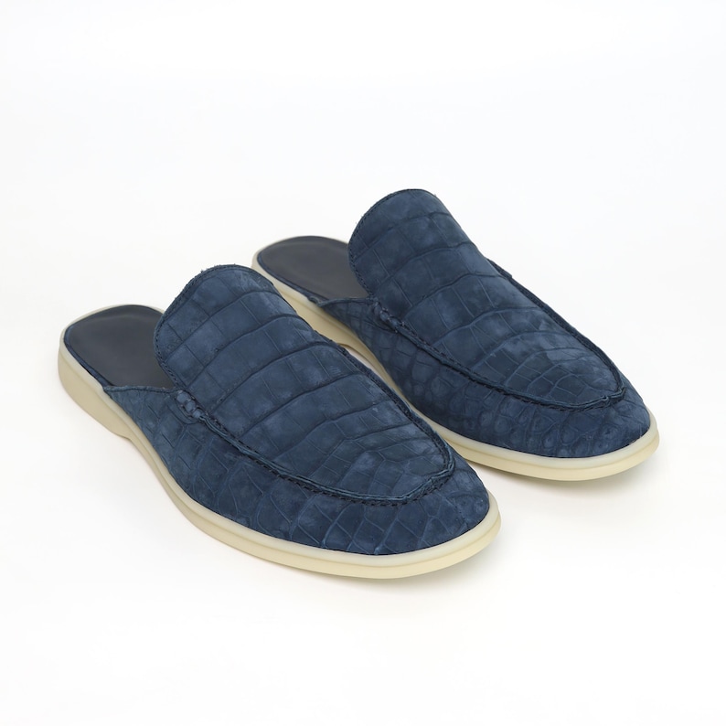 May include: A pair of blue suede slip-on shoes with a crocodile pattern. The shoes have a rounded toe and a cream-colored sole. The interior is a darker blue. The shoes are displayed on a white background.