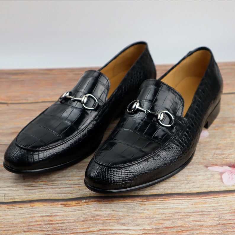 May include: Black leather loafers with a crocodile pattern and silver metal buckles. The loafers have a pointed toe and a low heel.