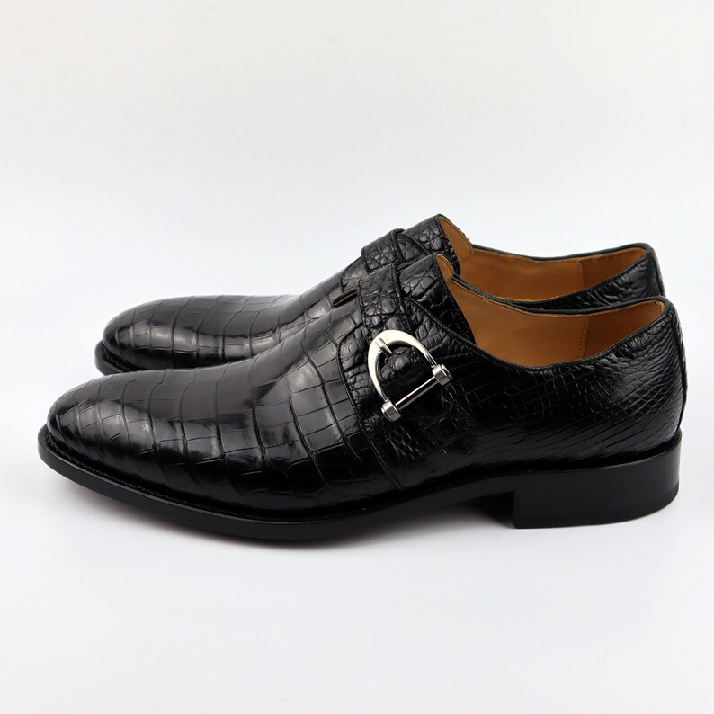 May include: Black leather dress shoes with a crocodile pattern and a silver buckle. The shoes have a pointed toe and a low heel.