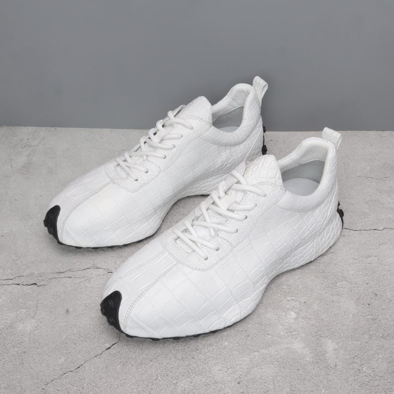 Puede incluir: Un par de zapatillas blancas con un patr&oacute;n texturizado similar al de un cocodrilo. Los zapatos tienen cordones blancos, un cuello acolchado y un acento negro en la puntera y el tal&oacute;n. Las zapatillas est&aacute;n dise&ntilde;adas para actividades deportivas o informales.