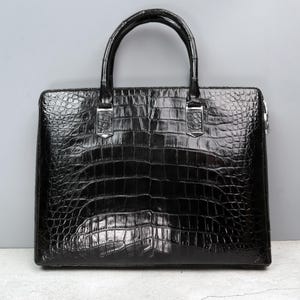 Pode incluir: Uma pasta de couro de crocodilo preta com ferragens prateadas. A pasta tem uma alça superior e um fecho de correr.