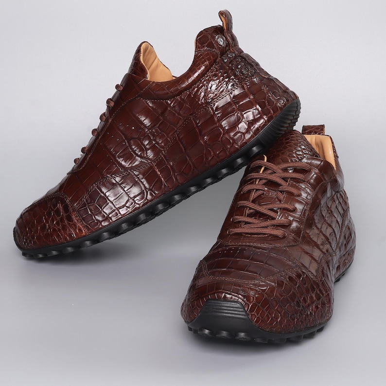 Puede incluir: Dos zapatillas de cuero marr&oacute;n con textura de piel de cocodrilo. Los zapatos tienen cordones marrones, suelas negras y un dise&ntilde;o de corte bajo. Se muestran sobre un fondo gris claro.