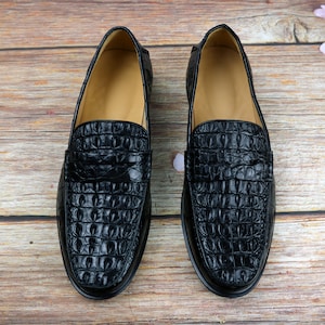 May include: Pair of black leather loafers with a crocodile skin texture. The shoes have a classic design with a rounded toe and a low heel. The interior is a light tan color.