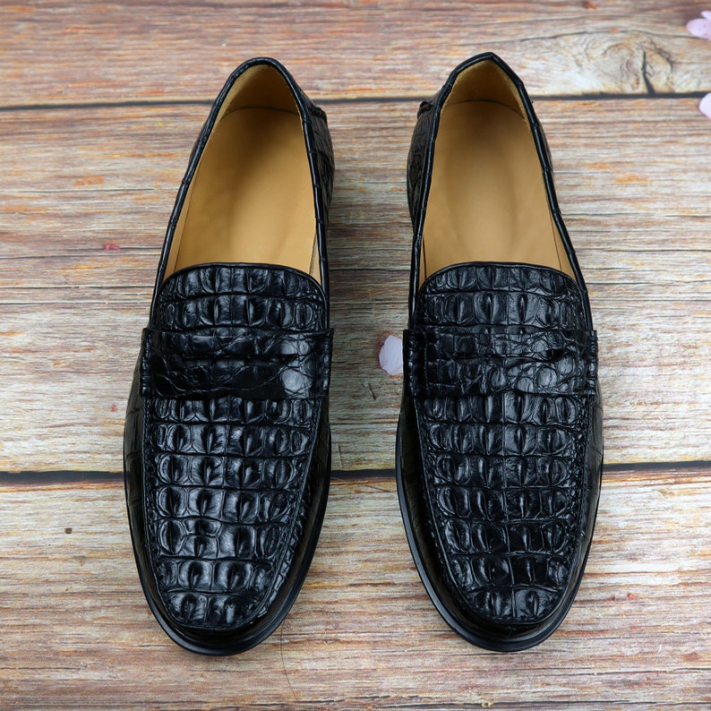 May include: Pair of black leather loafers with a crocodile skin texture. The shoes have a classic design with a rounded toe and a low heel. The interior is a light tan color.