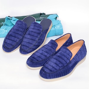 May include: A set of blue suede loafers with a crocodile pattern. The shoes have a rounded toe and a light-coloured sole. Two additional pairs of blue suede loafers are visible, along with rolls of blue and green leather in the background.