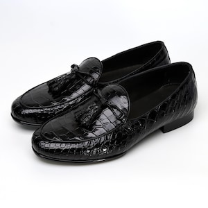May include: A pair of black leather loafers with a glossy finish. The shoes feature a crocodile skin texture and decorative tassels. The footwear is designed for a formal or semi-formal setting.
