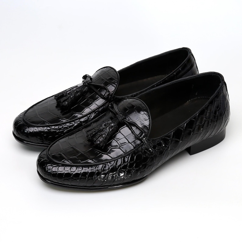 May include: A pair of black leather loafers with a glossy finish. The shoes feature a crocodile skin texture and decorative tassels. The footwear is designed for a formal or semi-formal setting.