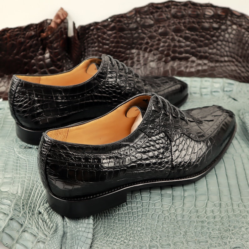 Op de afbeelding: Twee paar zwarte lakschoenen van alligatorshuid. De schoenen hebben een vetersluiting en een spitse neus. De schoenen liggen op een stukje alligatorshuid.