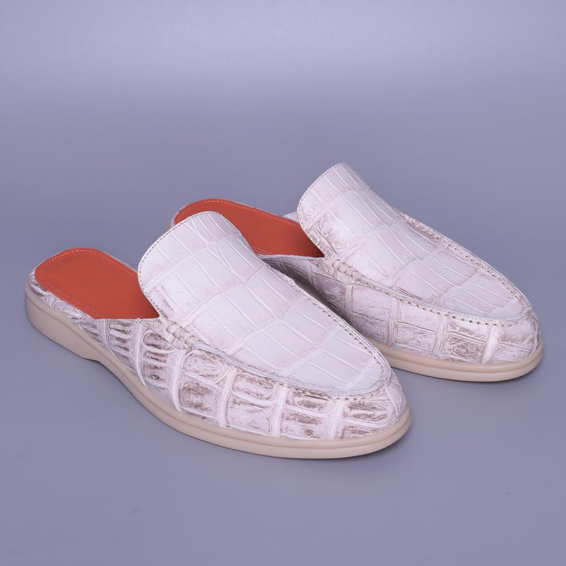 May include: A pair of white leather slip-on shoes with a crocodile skin texture. The shoes have a rounded toe and a tan sole. The interior is lined with orange leather. The shoes are displayed on a gray background.