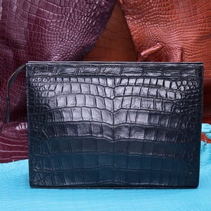 Puede incluir: Un bolso de mano rectangular de color negro con una textura prominente de piel de cocodrilo y acabado brillante. El bolso tiene un cierre de cremallera superior y está colocado frente a otras piezas de cuero texturizado en tonos morado, marrón y azul.
