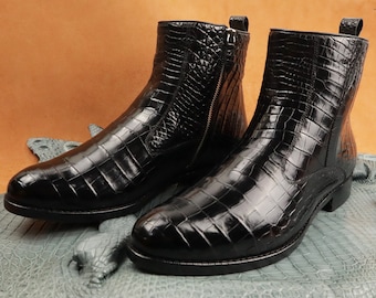 Pode incluir: Um par de botas de tornozelo pretas de couro de crocodilo com zíperes laterais. As botas têm bico fino e salto baixo.