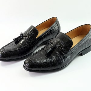 May include: Black leather loafers with a crocodile pattern and tassel detail. The loafers feature a metal buckle accent and a pointed toe.