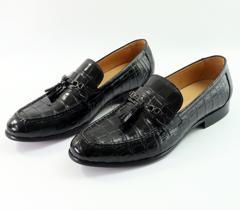May include: Black leather loafers with a crocodile pattern and tassel detail. The loafers feature a metal buckle accent and a pointed toe.