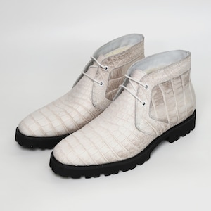 May include: A pair of off-white chukka boots with a crocodile skin texture. The boots have a lace-up design with white laces and black soles. The boots are made of a light-colored material.