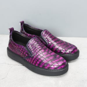 May include: A pair of slip-on shoes with a purple, textured, crocodile-like pattern. The shoes have a black sole and a small pull tab at the heel. The design features a dark purple elastic band on the top.