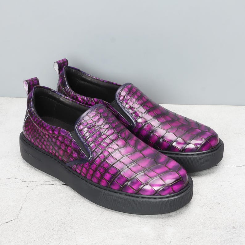 May include: A pair of slip-on shoes with a purple, textured, crocodile-like pattern. The shoes have a black sole and a small pull tab at the heel. The design features a dark purple elastic band on the top.