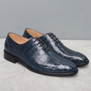 May include: A pair of navy blue leather dress shoes with a textured, crocodile-like pattern. The shoes have black laces and soles. The interior is a light tan colour. The shoes are displayed on a grey surface.