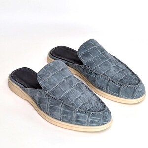 May include: A pair of blue suede leather slip-on shoes with a crocodile pattern. The shoes have a rounded toe, a black interior, and a cream-colored sole. The stitching is visible around the edges of the shoe.