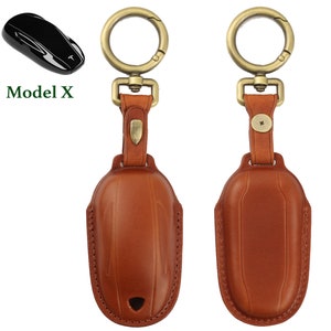 May include: A brown leather key chain with a gold metal key ring and a gold metal clasp. The key chain is designed to hold a Tesla Model X key fob. The key chain is shown next to a black Tesla Model X key fob. The text "Model X" is shown in green letters.